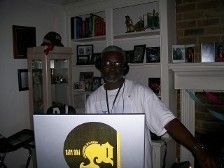 Older Black man wearing headphones, DJing with a laptop in a home setting.