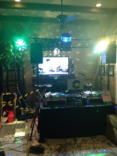 DJ setup with lights and equipment, including a table, speakers, and a TV. Green and white lighting.