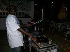 DJ at an outdoor event, operating equipment, wearing a white t-shirt and headphones, night setting.
