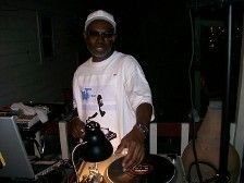 DJ wearing white shirt and cap, spinning records outdoors at night.