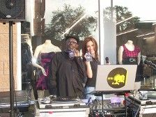 DJ duo at an outdoor event. One man and woman, holding purple CDs, set up with sound equipment in front of a store.