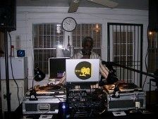 DJ in a room with sound equipment, laptop with logo, clock on wall, and white wall.