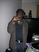 Man in glasses and hat holds a CD near a DJ setup. He looks up, brown striped jacket.