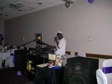 DJ at an event wearing white clothing and hat. The DJ is behind a sound system, set in a room.