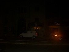 A dark street at night with a white van and headlights of a vehicle visible.