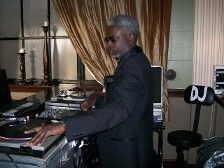 DJ at a gig, mixing music, wearing a dark suit, gray hair, indoor setting.
