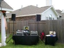 DJ setup outdoors with speakers, table, and equipment in front of a wooden fence and house.