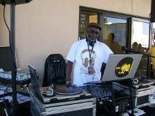 DJ standing at equipment outside, wearing hat and t-shirt. Laptop and turntable visible.