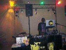 DJ at event, mixing music.  Black table, sound equipment, stage lights, person standing, laptop.