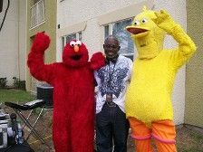 Man with Elmo and Big Bird costumes, smiling outside. Elmo raises arms.