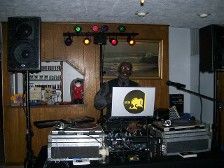 DJ in front of equipment with laptop, speakers, and lights, inside a room with art.