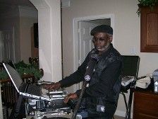 An older Black man wearing a hat, DJing with a laptop in a home setting.