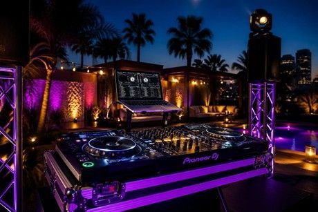 DJ turntable setup lit in purple at an outdoor night party with palm trees and city lights