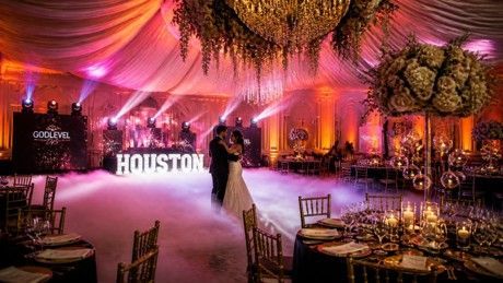 Elegant banquet hall with pink-orange lighting, round tables, and a stage backdrop reading “HOUSTON” with performers