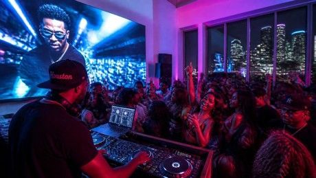 DJ performing for a packed nightclub crowd with blue and pink neon lighting and a large screen behind them