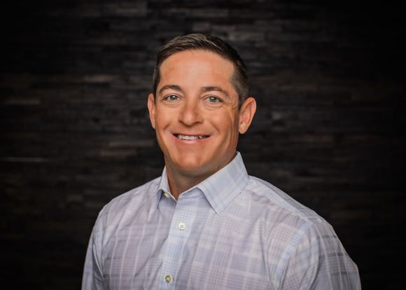 Man in a light blue button-down shirt smiles, looking directly at the camera against a dark wooden backdrop.