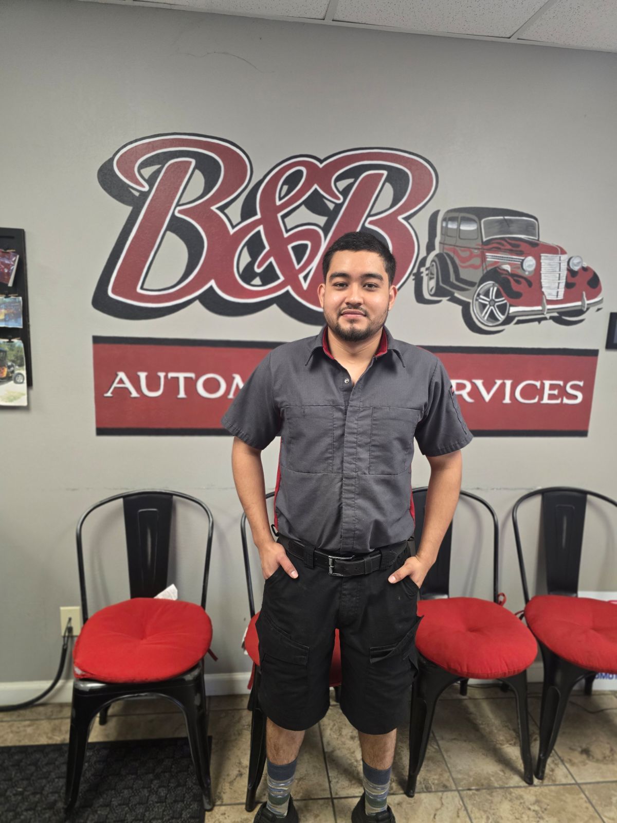 Man standing in front of a B&B Automotive Services sign with chairs and a red backdrop behind him.