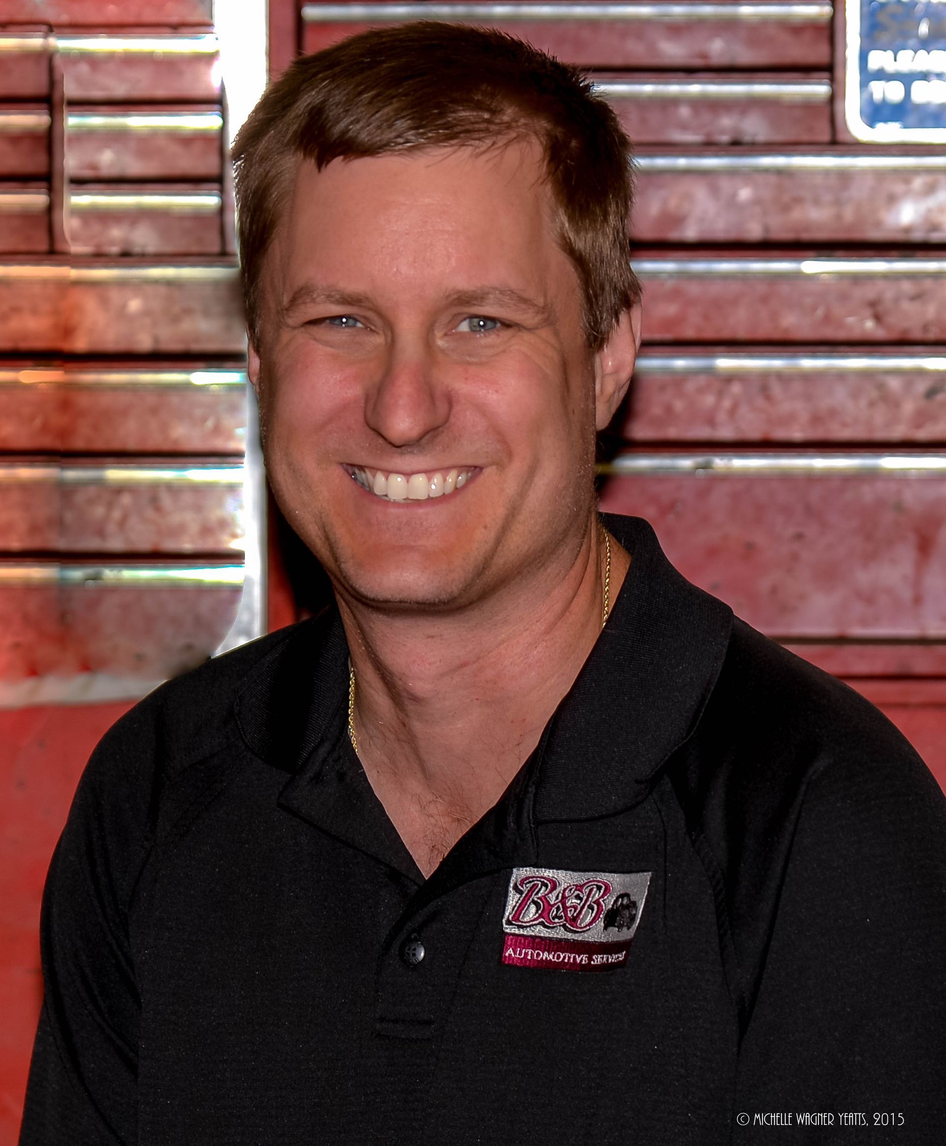 Smiling man in a black polo shirt standing before a wooden wall