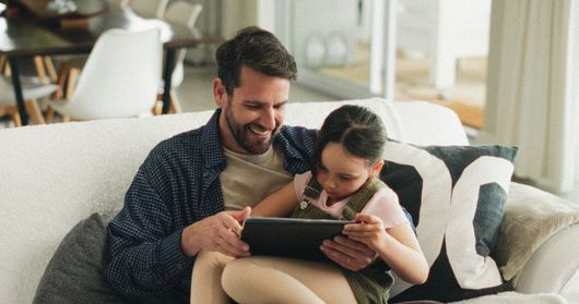 Deux personnes assises sur un canapé sourient et utilisent une tablette ensemble dans un salon lumineux.