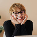 Personne souriante portant des lunettes et ayant les cheveux courts, le visage appuyé sur ses mains, sur fond beige.