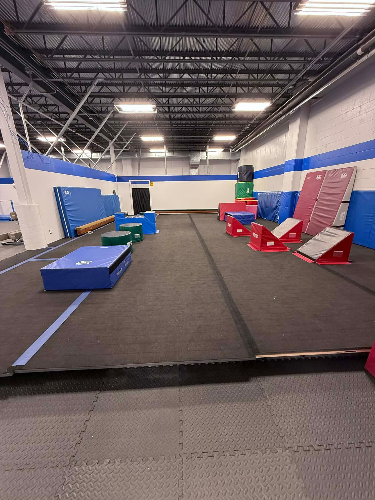 Indoor gym with padded obstacle course mats, ramps, and blocks under high industrial lights.