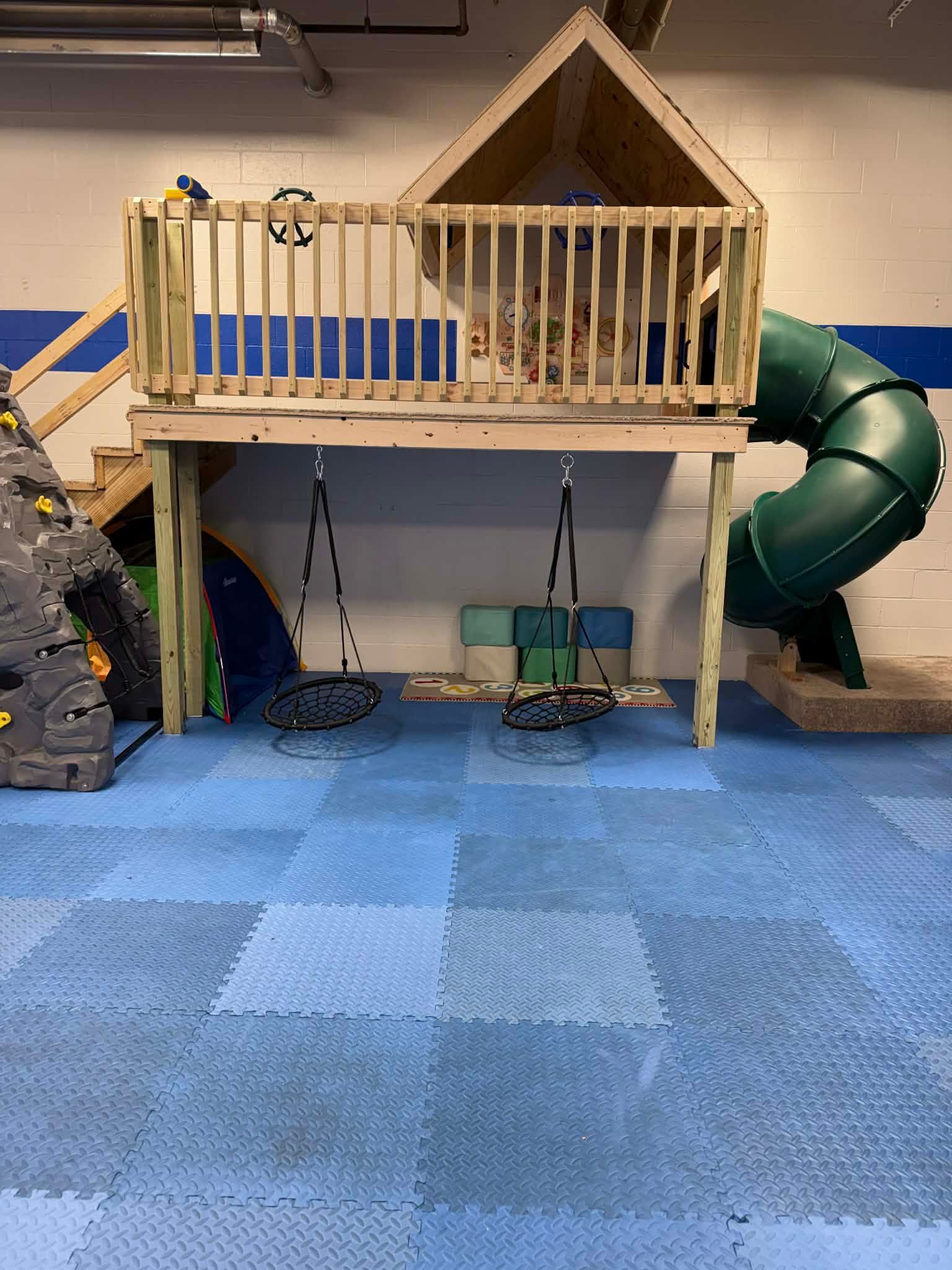 Wooden indoor play loft with railing, slide, and climbing area over blue foam floor mats