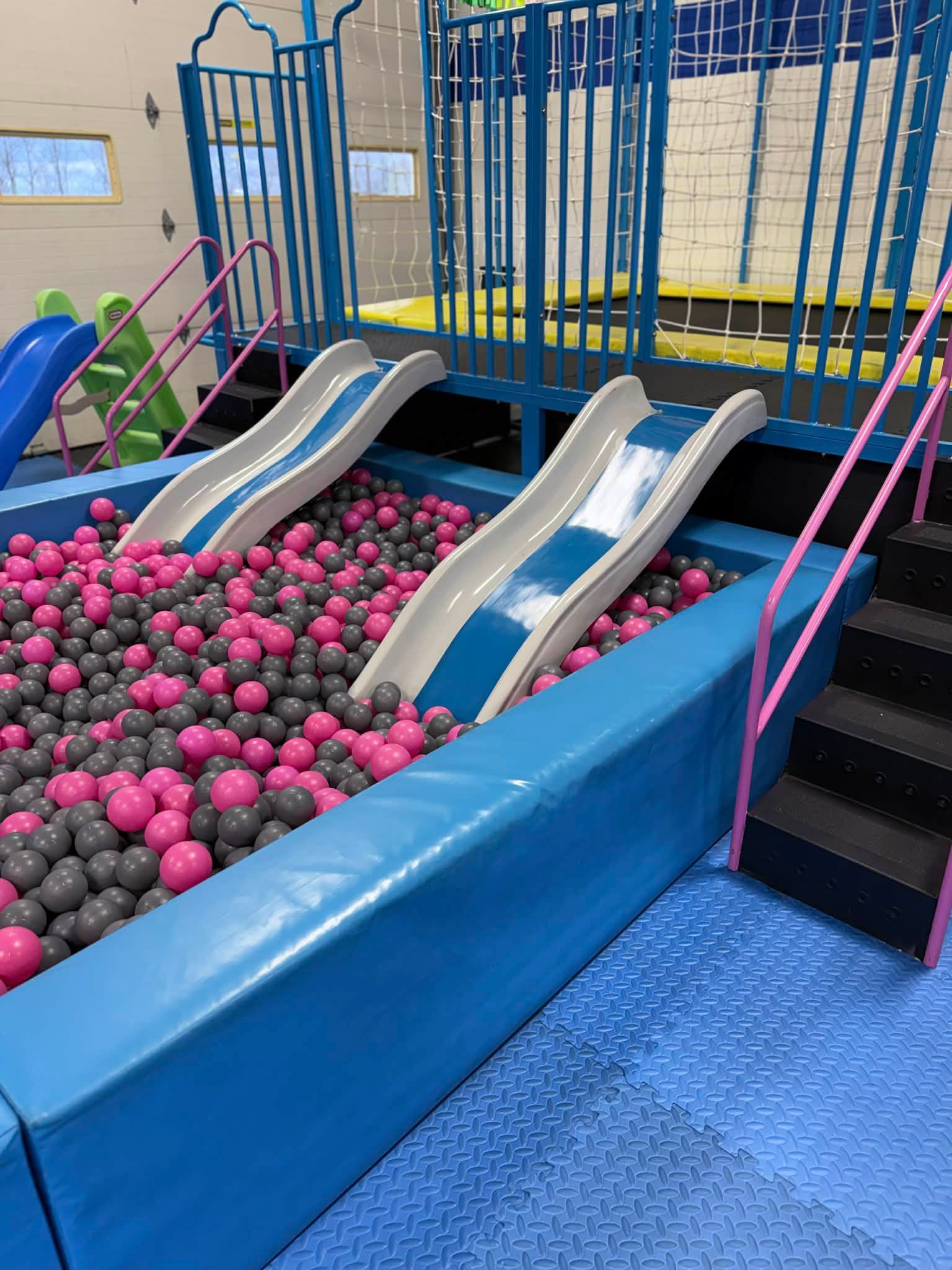 Indoor playground ball pit with pink and gray balls and two white slides beside blue railings.