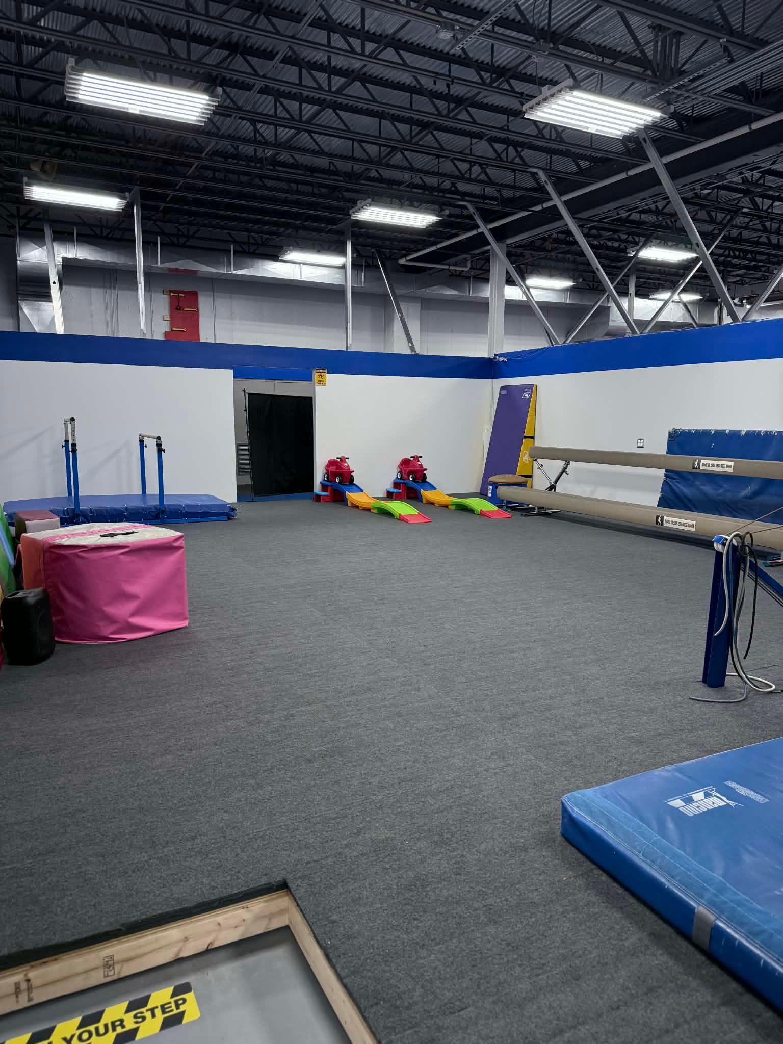 Indoor gymnastics training area with mats, balance beams, and colorful equipment on gray carpet under bright lights