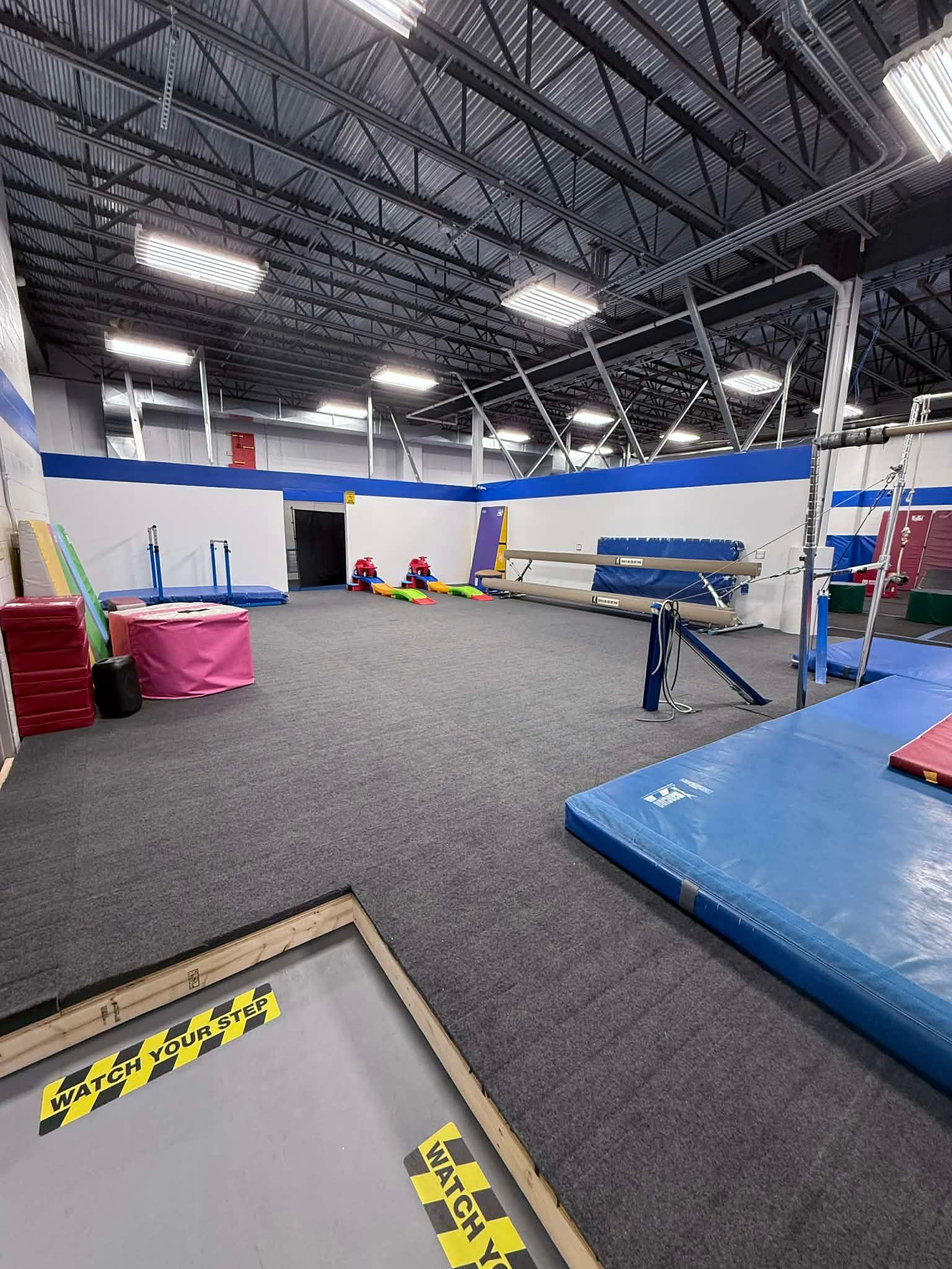 Indoor gymnastics gym with blue mats, balance beams, and tumbling equipment on a carpeted floor.
