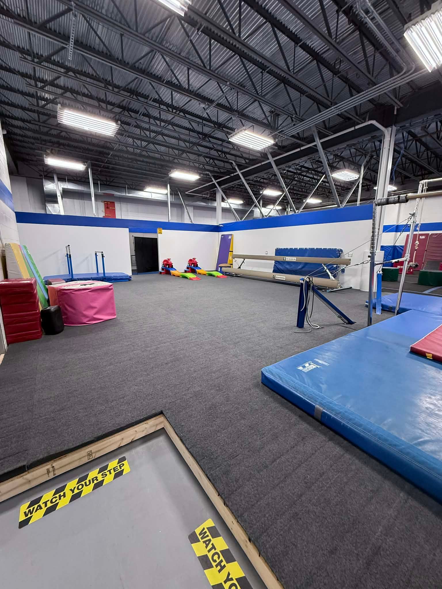 Indoor gymnastics gym with blue mats, spring floor, balance beam, and high ceiling lights