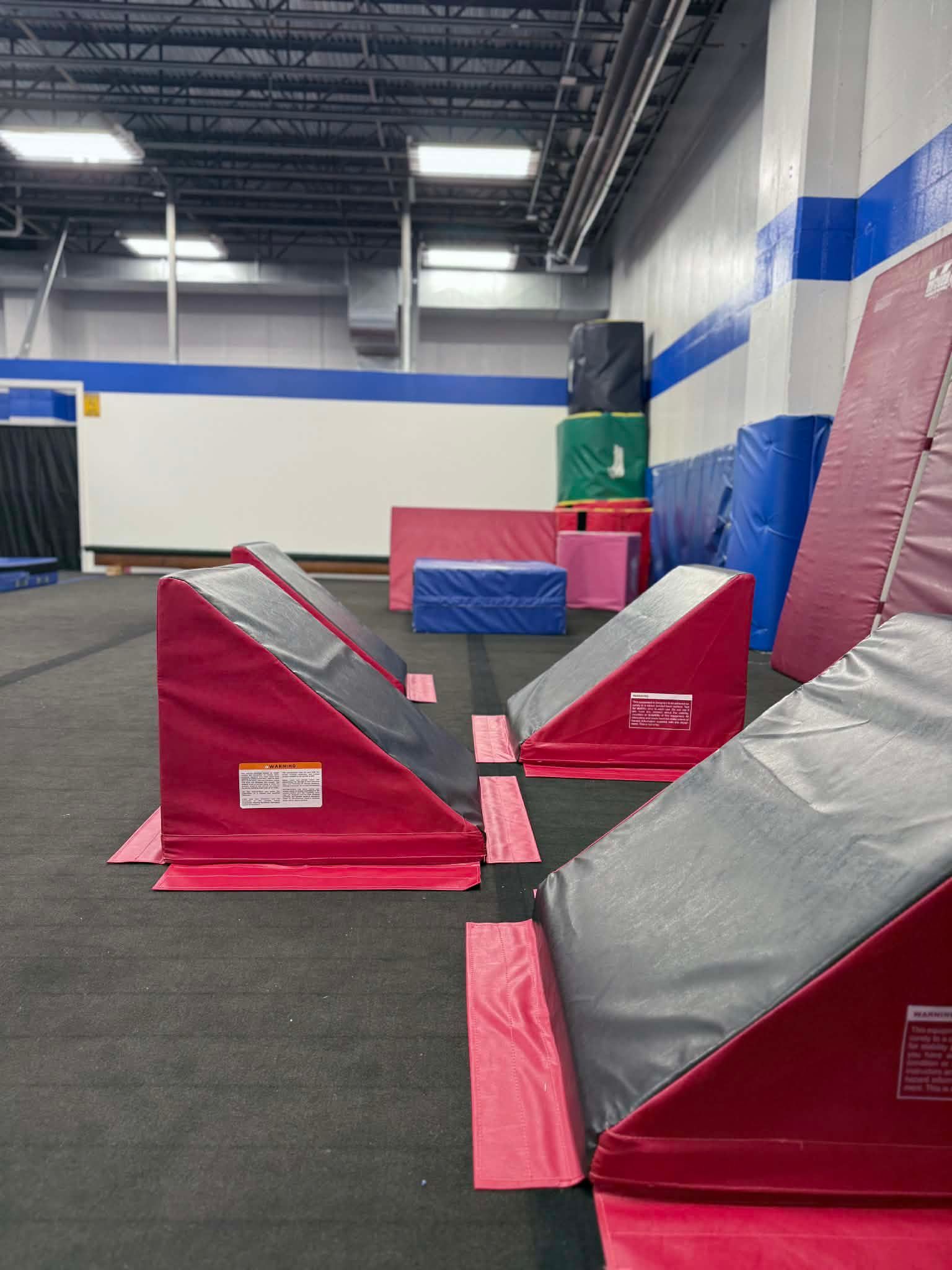 Indoor gym with red and gray foam ramps and mats on a black floor beneath high industrial ceilings.