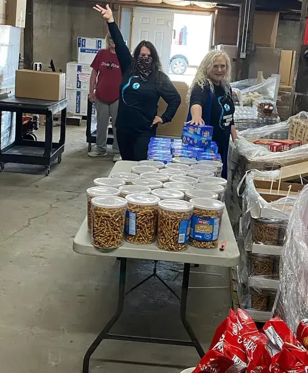 Two women are standing in front of a table filled with jars of pretzels.