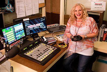 A woman is sitting at a desk in a radio station holding a microphone.