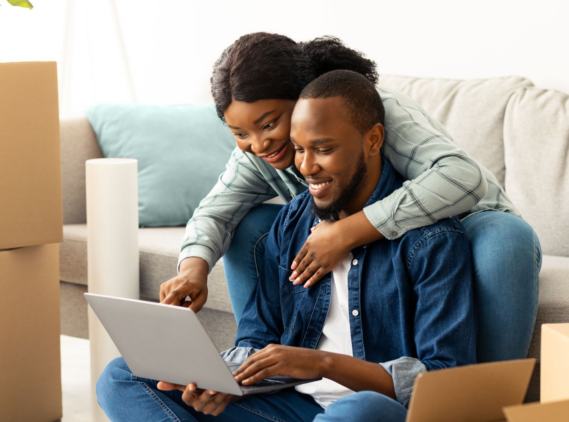 Two people smiling at a laptop on a couch with moving boxes nearby