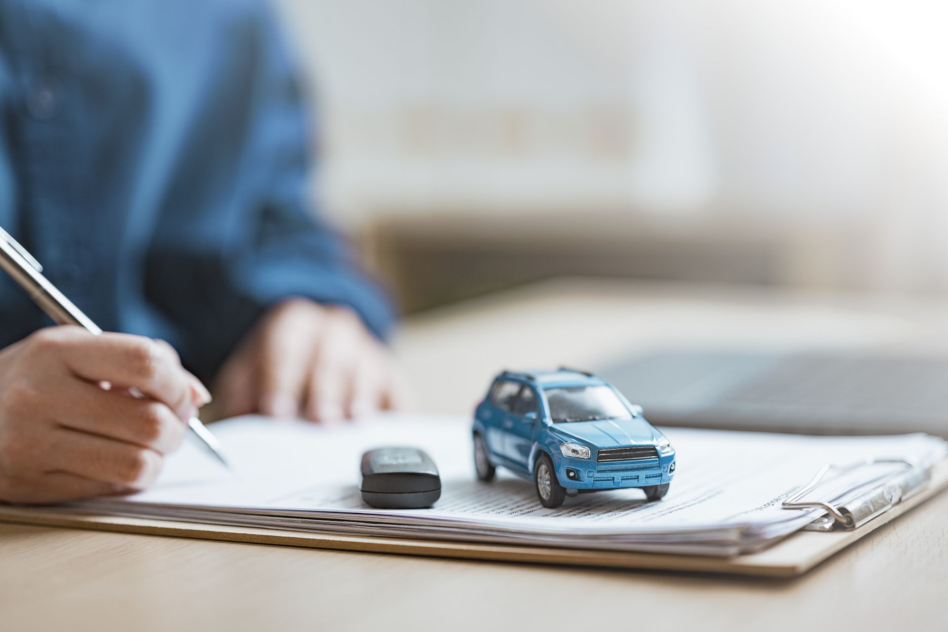 Hand writing on a clipboard beside a small blue toy car and a key, suggesting car insurance or inspection paperwork