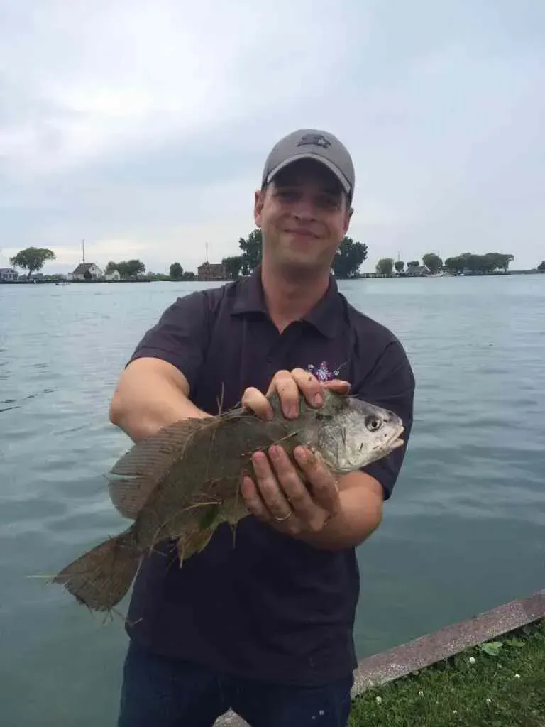 Man holding a fish by the water, wearing a hat and black shirt. Cloudy day.