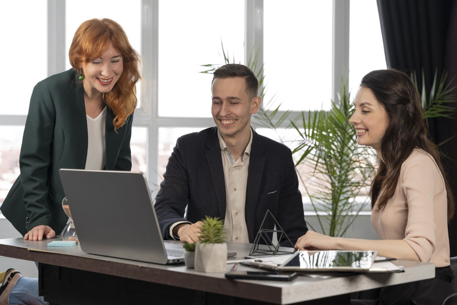 Three people smiling, looking at laptop screen in office, discussing, natural light.