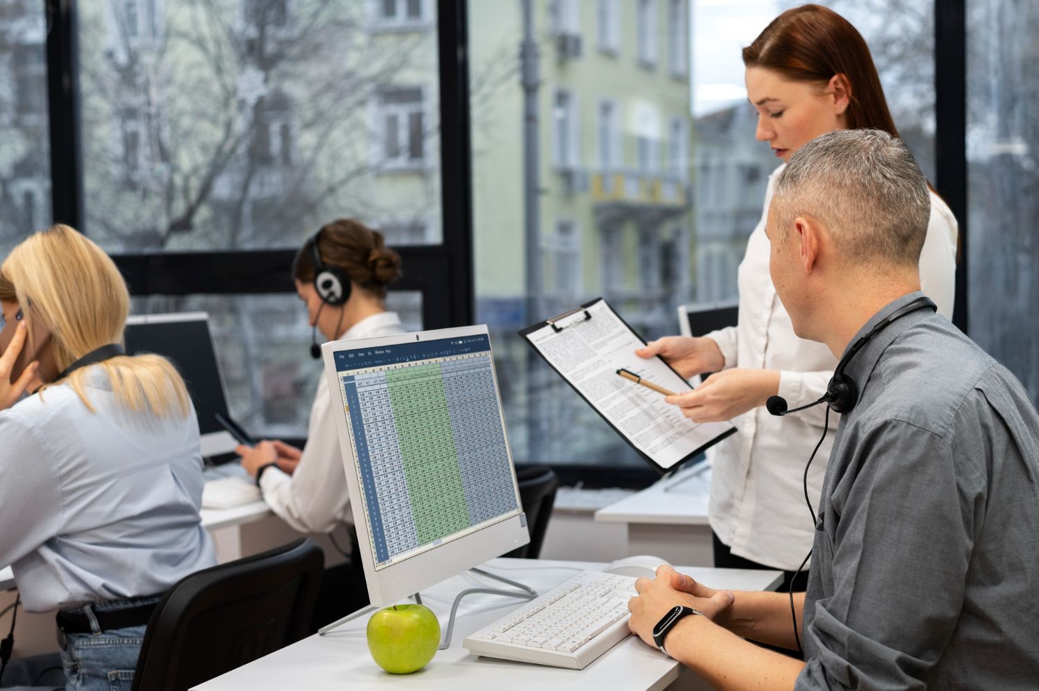 People working at computers in an office setting; a supervisor reviews paperwork with a colleague.