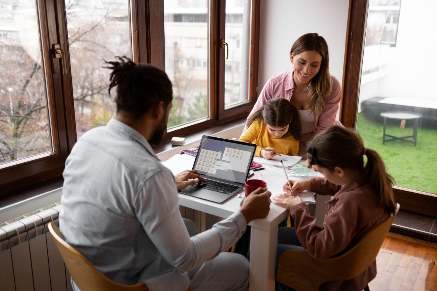 Family at table: person using laptop, children drawing, woman watching. Near windows, indoor setting.