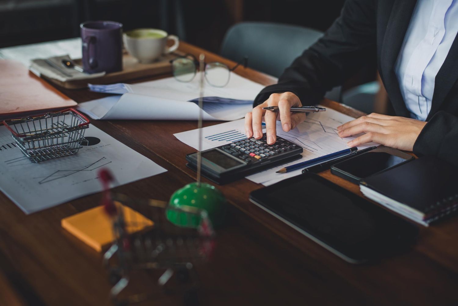 Person in suit uses calculator at a cluttered desk with papers, phone, and coffee cup.
