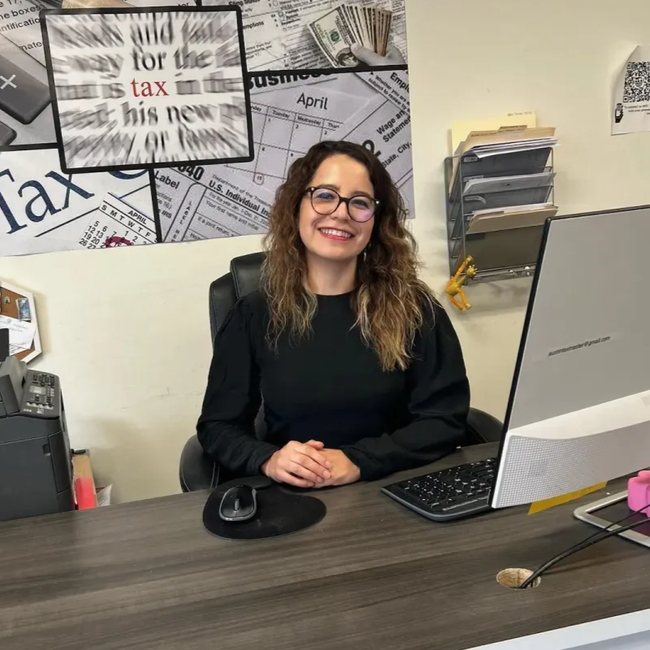 Woman at desk in office, smiling, with a computer and tax documents in the background.