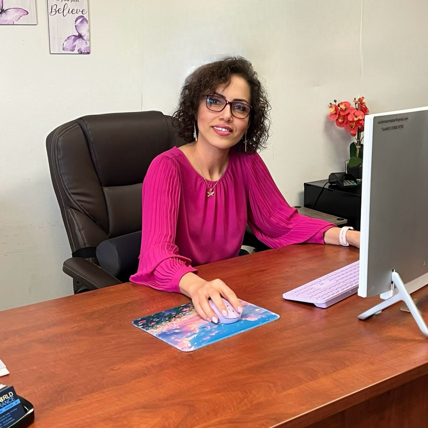 Woman sitting at a desk in an office, smiling, wearing a black blazer.