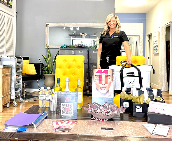 Woman in scrubs stands behind a table with products. Yellow accent chair, wine, and a cooler are visible.