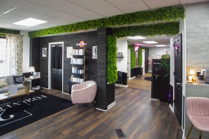 Two women conversing in a beauty salon reception area, with a desk, greenery wall, and a treatment room visible.