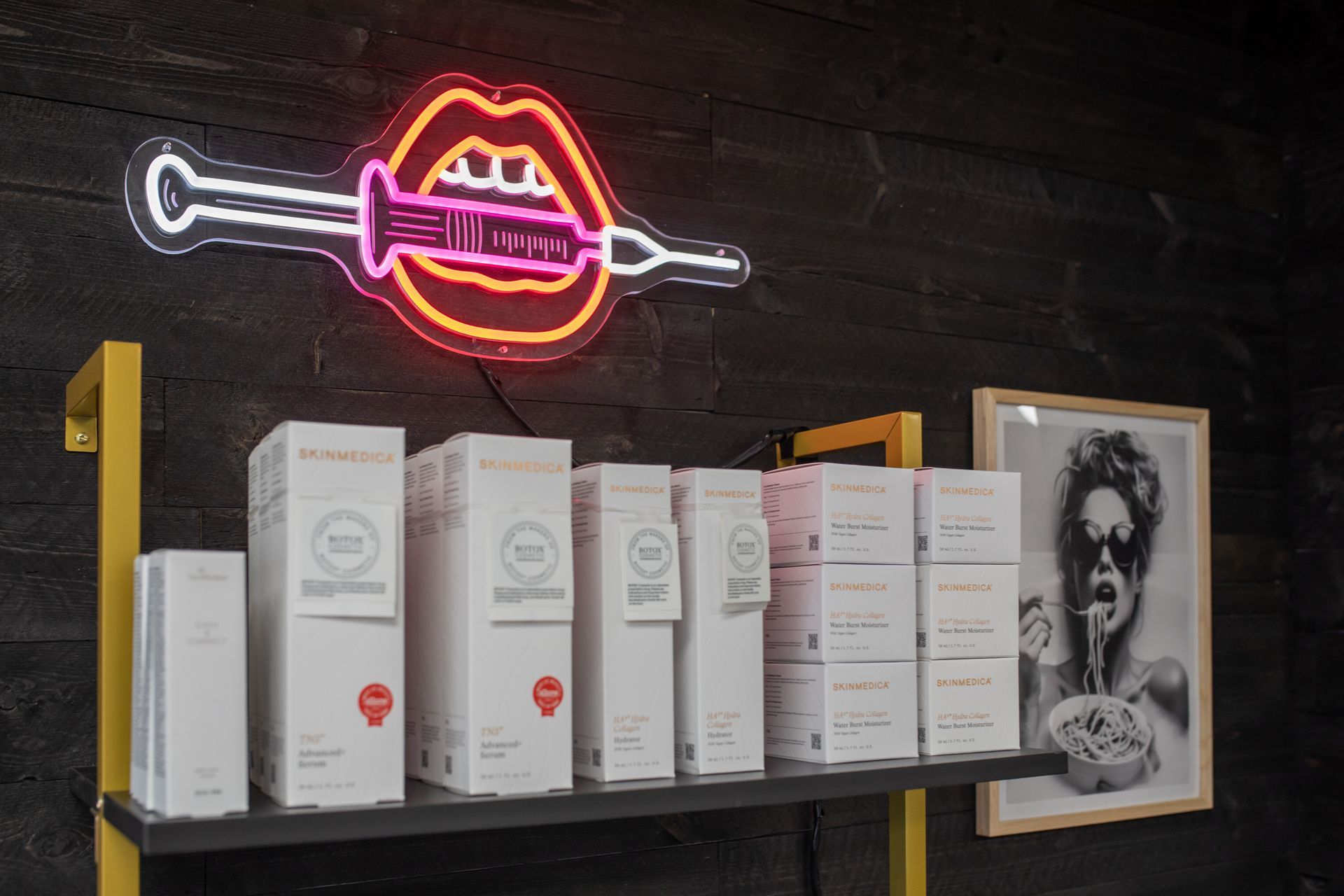 Shelf display with product boxes, brochures, and neon lips with syringe sign.