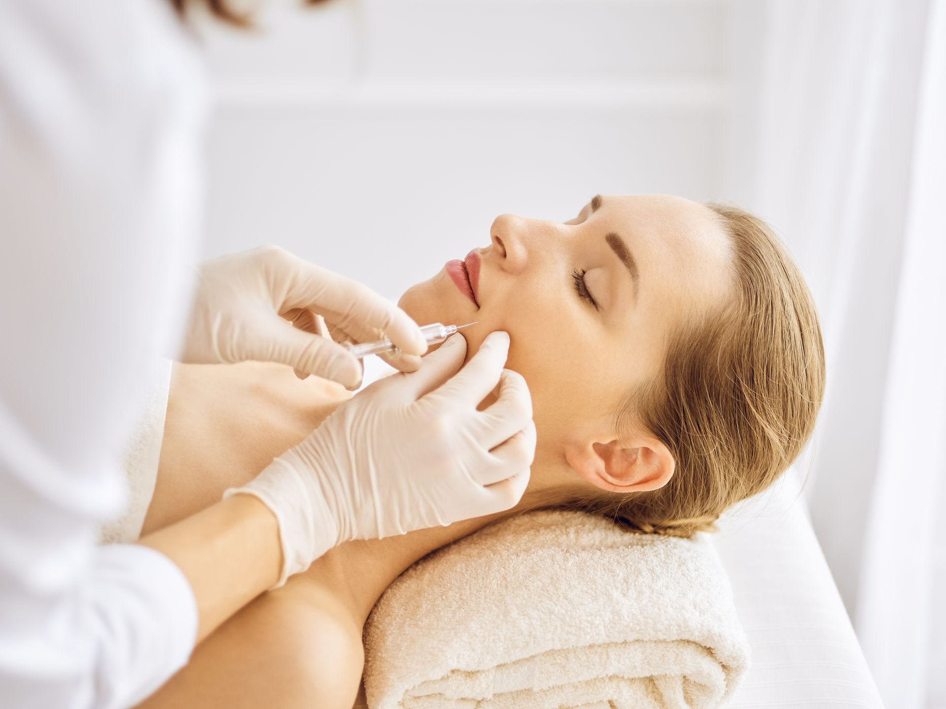 Woman receiving facial injection in a medical setting, white gloves, towel, closed eyes.