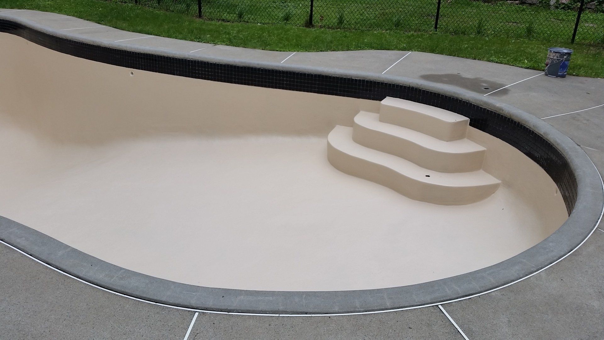 Piscine beige vide avec escalier, entourée d'une terrasse en béton et d'une clôture noire.