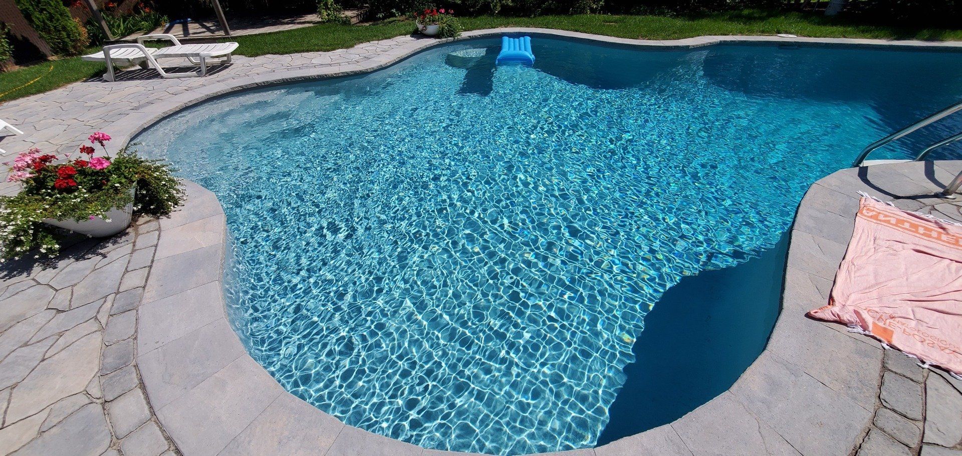 Piscine à l'eau bleue et terrasse en pierre. Des fleurs rouges dans une jardinière trônent au bord de la piscine.