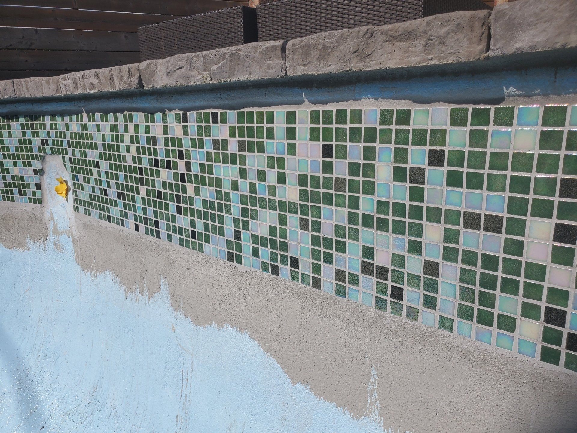 Bord de piscine, mosaïque aux tons verts et blancs, bordure en béton. Surface bleue visible.