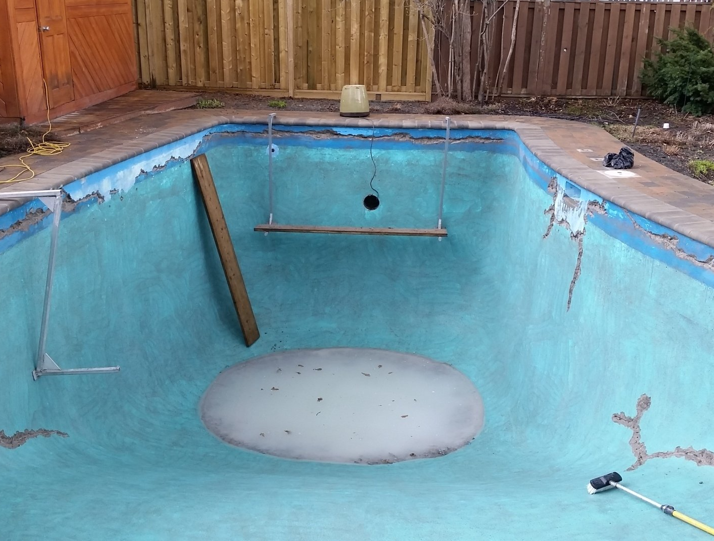 Piscine vide, vieillissante et bleue, aux parois fissurées et aux débris. Clôture en bois et outils visibles.